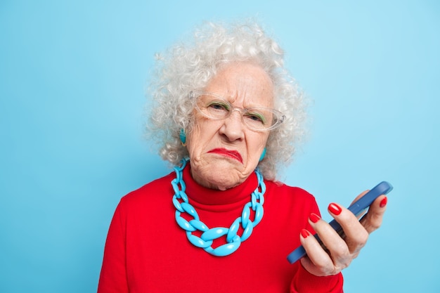 Photo of displeased wrinkled old curly woman holds mobile phone checks message frowns face wears spectacles red jumper and necklace