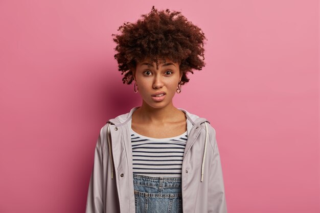 photo of displeased young curly woman smirks face, has negative reaction, hears nonsense, looks in dissatisfaction, dressed casually, poses against rosy wall. negative human expressions