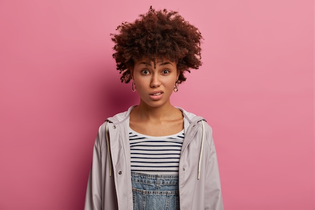 Photo of displeased young curly woman smirks face, has negative reaction, hears nonsense, looks in dissatisfaction, dressed casually, poses against rosy wall. Negative human expressions