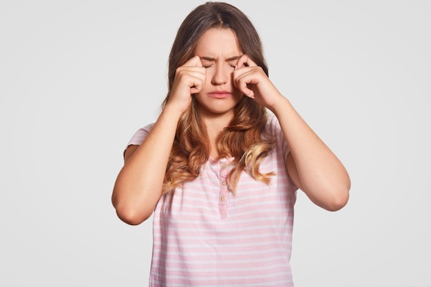 Free photo photo of displeased young girl rubs eyes, being in low spirit, wears casual domestic clothes, poses on white. woman had bad sleep at night. bedding concept. people and tiredness