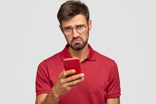 Photo of displeased young unshaven guy holds modern red smart phone, being unhappy to read neagtive news in internet, messages with friends in social networks, connected to wireless internet