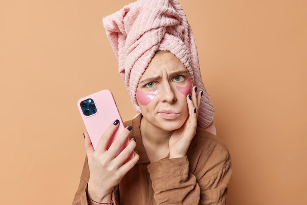 불쾌한 젊은 여성의 건강한 피부를 가진 사진은 잠옷을 입은 미용 하이드로겔 패치를 적용하고 목욕 후 머리에 수건을 착용하고 갈색 배경에 대해 휴대폰 포즈를 사용합니다