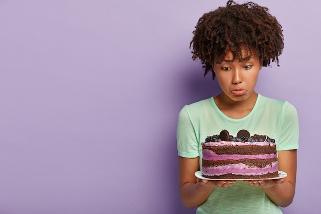 Free photo photo of dissatisfied afro american woman holds plate of blueberry sweet cake, purses lower lip, has no good will, wants to eat delicious dessert but keeps to diet