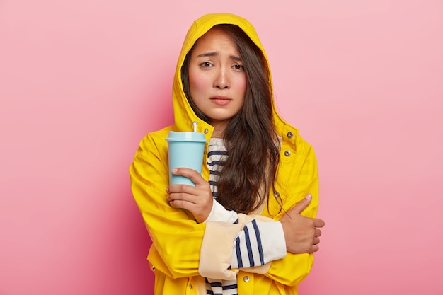 不満を持ったアジア人女性の写真は、顔を笑い、腕を組んで、雨の中を歩いた後の寒さから震え、黄色いレインコートを着て、持ち帰り用のコーヒーを飲み、温かい飲み物で体を温めます