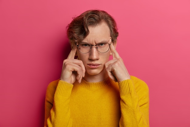 Photo of dissatisfied man suffers from headache, frowns face, touches temples with fingers, tries to focus, remembers something, wears spectacles, yellow sweater, isolated on pink wall
