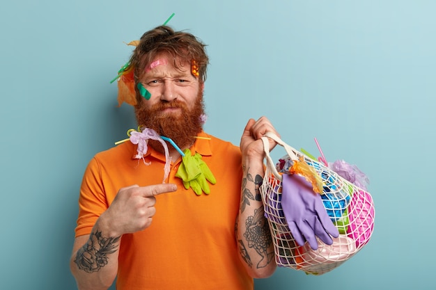 Photo of dissatisfied red haired man with thick bristle, points fore finger at bag filled of plastic rubbish, wears casual orange t shirt, has tattooed arm, stands over blue wall. Earth Day