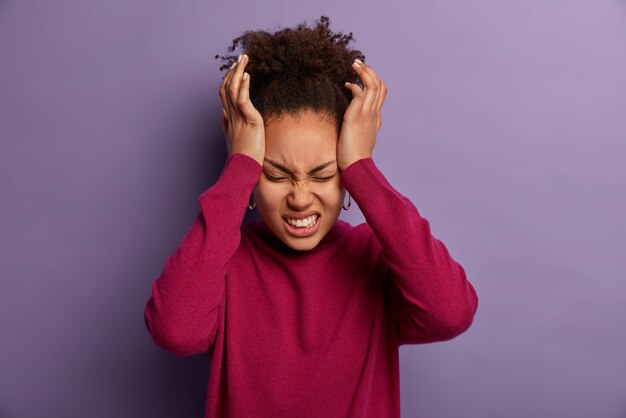 Photo of dissatisfied woman keeps hands on temple, suffers from unbearable headache, clenches teeth with pain, being exhausted after work, wears burgundy turtleneck, isolated on purple wall.