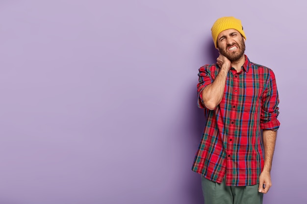 Photo of dissatisfied young man with bristle, feels stiffness in neck, tilts head, clenches teeth, dressed in checkered shirt, stands against purple background blank space for your promotional content