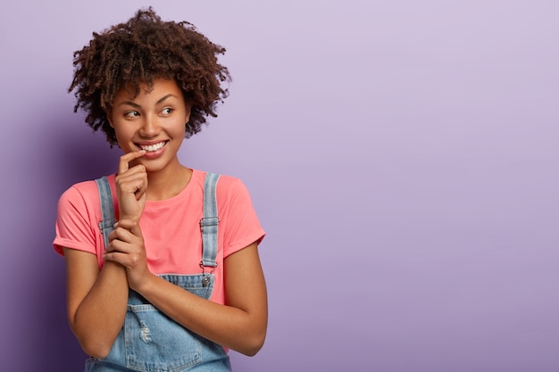 Photo of dreamy romantic woman keeps index finger near lips, has pleased thoughtful expression, has Afro hairstyle, wears pink t shirt and jean sarafan