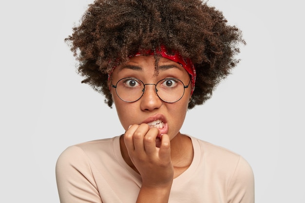 Photo of embarrassed dark skinned woman looks anxiously, bites finger nails