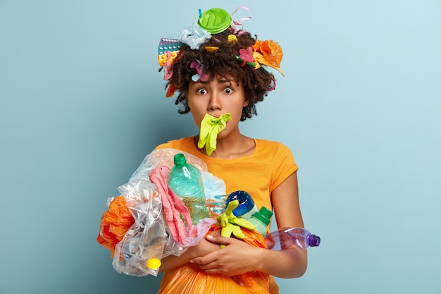 Photo of embarrassed young curly Afro American woman has rubber glove in mouth, carries plastic garbage, worried by global environmental pollution, isolated on blue wall. Ecology concept