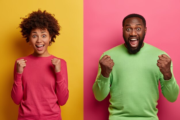 Photo of emotional dark skinned female and male clench fists, exclaim and support favorite football team, have overjoyed face expressions, dressed in casual wear, isolated on yellow and pink wall