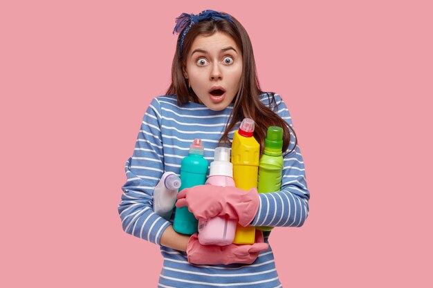 Photo of emotional European housemaid opens mouth widely, stares in stupor, carries bottles of cleaning supplies, cares of sanitary and hygiene