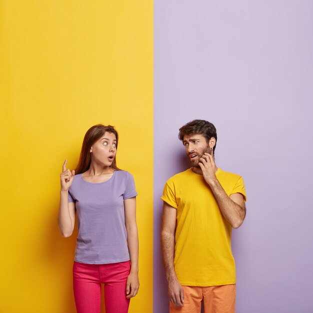 Photo of emotional impressed woman tries to explain something to man, points above with shocked expression, displeased guy scratches bristle, wears yellow t shirt