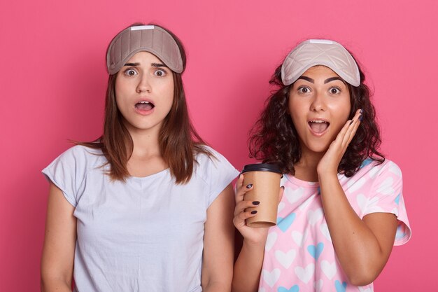 Photo of emotional pretty young females looking directly  holding coffee, openingand eyes mouth widely, being shocked