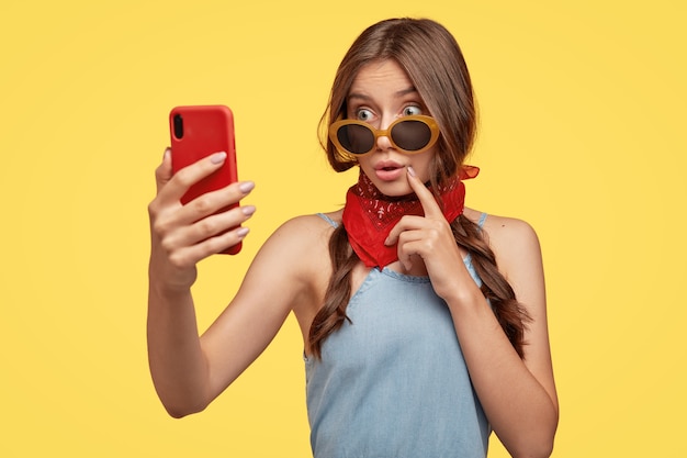 Photo of emotive astonished wondered woman looks with amazement through sunglasses, holds modern cell phone, takes picture of herself, shocked with new appearance, wears red bandana around neck