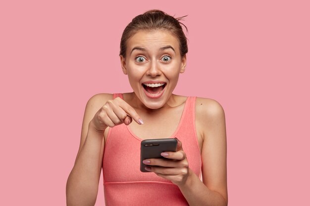 Фотография эмоциональной кавказской спортивной женщины с удивленным выражением лица, показывает указательным пальцем на мониторе сотовой связи, носит повседневный жилет, получает хорошее уведомление, изолирована на розовой стене