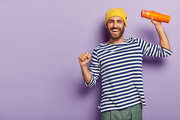 Photo of energized cheerful man tourist dances playfully, dressed in fashionable clothes, carries thermos, smiles gladfully, has happy mood, isolated over purple background.