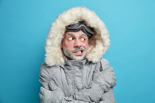 Photo of European man trembles from cold after going skateboarding crosses hands over body tries to warm himself wears grey winter jacker with fur hood and gloves has frozen face covered by ice