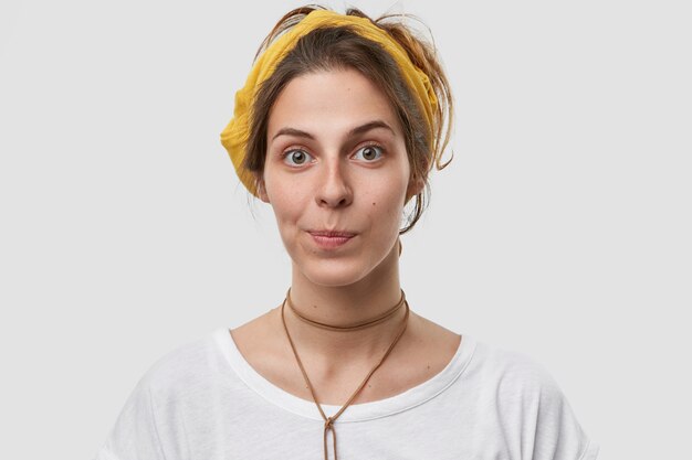 Photo of European woman with attractive appearance, purses lips, has healthy soft skin, wears yellow headband, casual t shirt