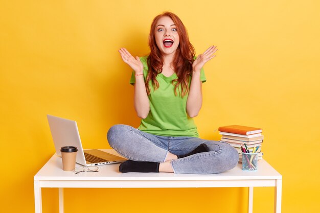 Photo of excited student girl sitting at desk surrounded with exercise books, lap top, coffee, keeping mouth widely opened spreading palms aside, isolated over yellow background.