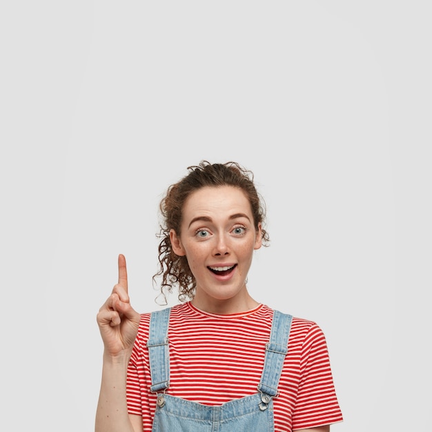 Photo of excited surprised woman with freckled skin, points with index finger, shows something amazing upwards