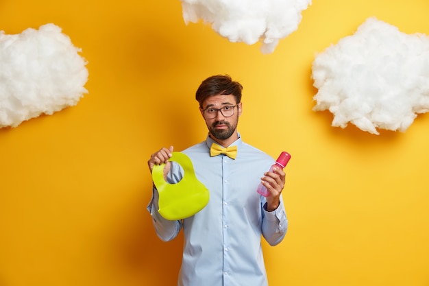 Photo of exhausted bearded father going to feed baby, holds bottle and bib, cares of newborn, dressed formally, poses against yellow  with white clouds. Fatherhood, parenthood concept