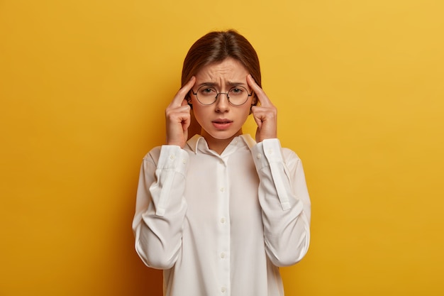 Photo of exhausted European woman holds fingers on temples, suffers from headache, thinks hard, smirks face, dressed in white elegant shirt, gets feelings together, ponders important decision