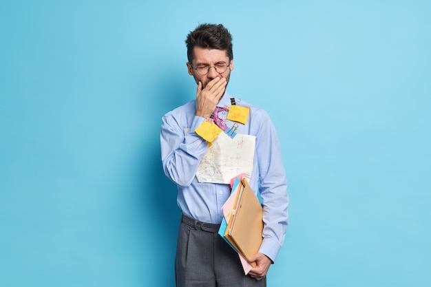 Фотография измученного мужчины зевает после долгих часов работы, готовит финансовый отчет, носит официальную одежду, позирует в помещении