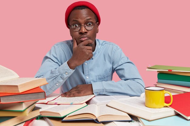 Photo of fashionable hipster in red hat, keeps hand on chin, looks surprisingly at camera, has much work before session, reads textbooks for education