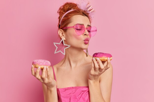 Foto di donna europea glamour rossa alla moda tiene le labbra piegate tiene due ciambelle appetitose vuole mangiare dessert dolce