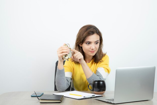 Фото женского офисного работника, сидящего за столом и указывающего на будильник. Фото высокого качества