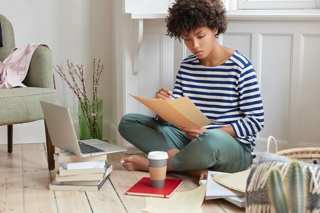 Photo of financial manager creats report in cozy place, sits crossed legs near opened laptop, holds papers, develops strategy for making business