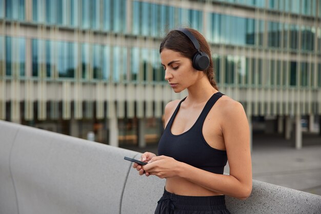 Photo of fit female model chooses song for workout downloads song for her running playlist has break after training wears sportswear poses in modern setting