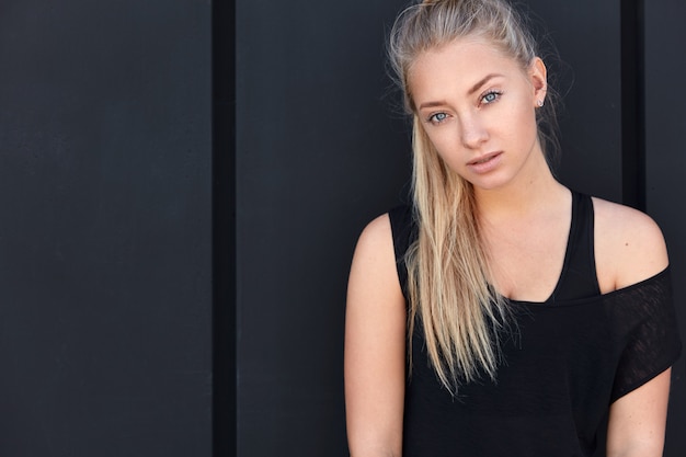 Photo of fitness blonde female with pony tail, dressed casually, has rest after cardio training, looks seriously, poses against black wall