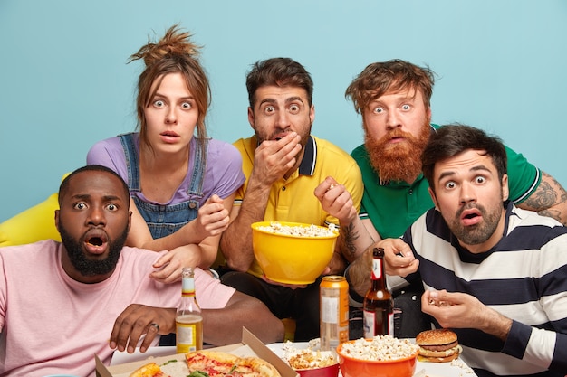 Free photo photo of five mixed race woman and men watch thriller movie, horrible news, look in panic, eat popcorn, stare with bugged eyes, isolated over blue wall, being scared. scary film at home