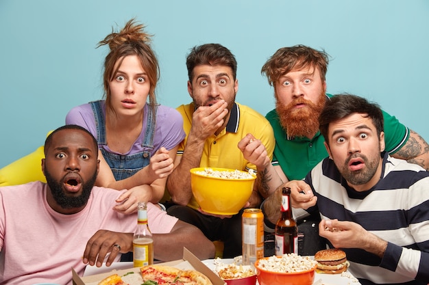 Photo of five mixed race woman and men watch thriller movie, horrible news, look in panic, eat popcorn, stare with bugged eyes, isolated over blue wall, being scared. Scary film at home