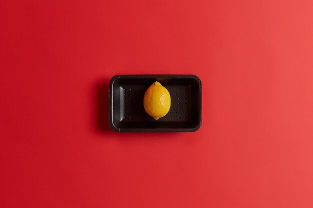 Photo of fresh ripe yellow single lemon on black tray bought in supermarket isolated over red background. Whole exotic fruit containing much vitamin C. Ingredient for cooking cold summer lemonade