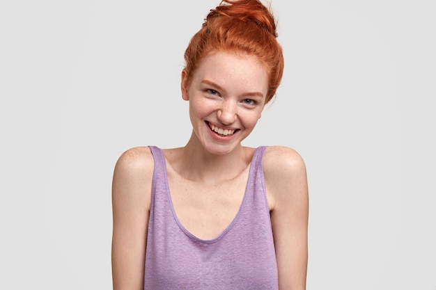 Photo of friendly sociable woman has red hair and perfect freckled skin, gazes happily, has tender smile, wears casual oversized vest, shows bare shoulders, models against white wall