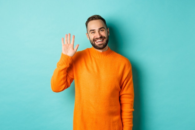 Free photo photo of friendly young man saying hello, smiling and waiving hand, greeting you, standing in orange sweater over light turquoise wall.