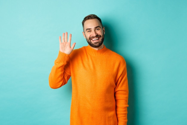 Photo of friendly young man saying hello, smiling and waiving hand, greeting you, standing in orange sweater over light turquoise wall.
