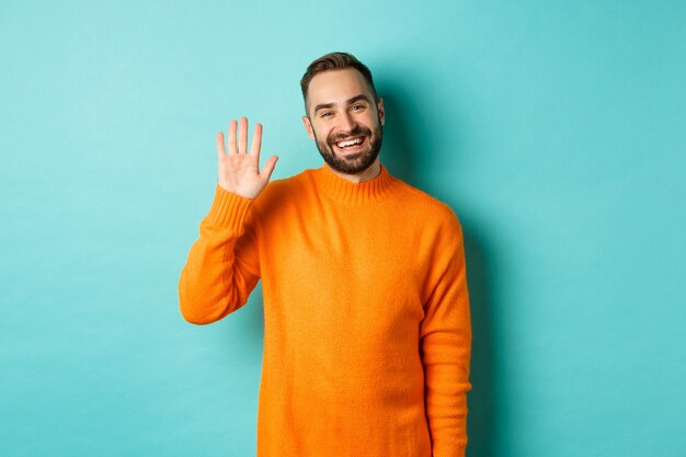 밝은 청록색 벽 위에 주황색 스웨터에 서서 인사하고, 인사하고, 손을 흔드는 친절 한 젊은 남자의 사진.