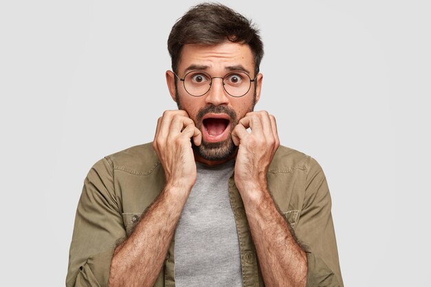 Photo of frightened emotive bearded young male keeps jaw dropped, expresses fear and disbelief, keeps hands near opened mouth, stares with anxious look, isolated over white wall