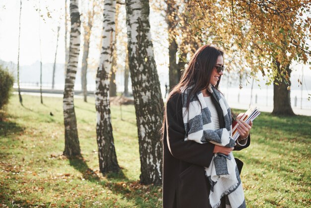 Photo from the side. Young smiling brunette in sunglasses stands in the park near the trees and holds notepad