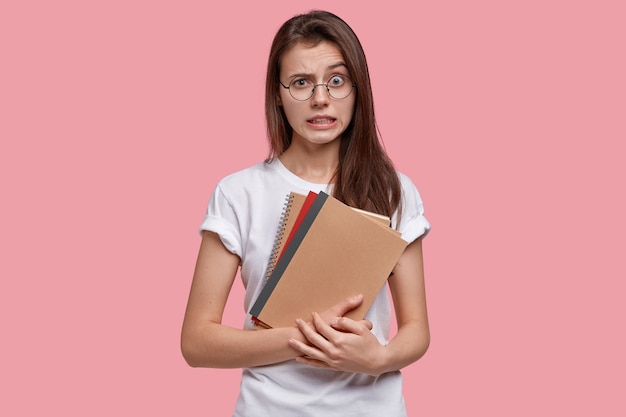 Photo of frustrated puzzled young female looks in bewilderment, raises eyebrow, has dark hair, white casual t shirt