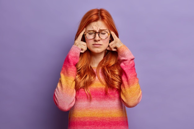 Free photo photo of frustrated redhead young woman keeps fingers on temples suffers headache or severe migraine closes eyes to reveal pain wears spectacles and jumper.