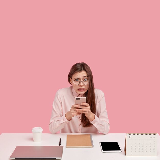 Photo of frustrated woman holds mobile phone, sends money via online bannking app, surrounded with laptop computer, spiral notepad, calendar