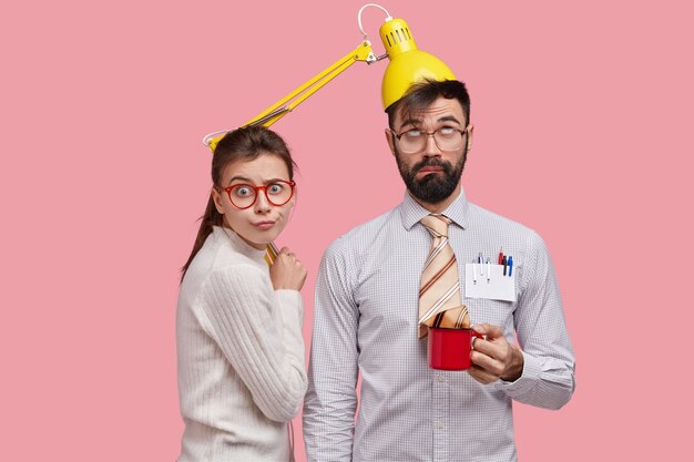 Photo of funny bearded young man has lamp on head, tie in cup of tea, dressed in formal clothes, his female partner stands near