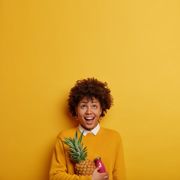 Photo of funny ethnic woman concentrated upwards, holds fresh pineapple and smoothie, has healthy nutrition, dressed in casual wear, isolated over bright yellow wall, copy space for your text