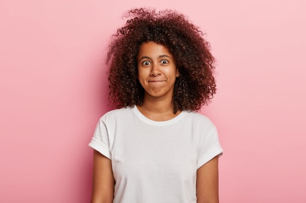 面白い女性の写真は、巻き毛の太い髪、唇を一緒に押し、幸せそうな顔をしていて、白いTシャツを着て、ピンクの壁に隔離されています。格好良い若いアフリカ系アメリカ人の女の子は幸せを表現します。